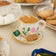 Hand-felted ornament of a floral teacup with a matching saucer, featuring embroidered golden rims, a tiny felt tea bag, and two sugar biscuits on the side. Crafted from merino-blend wool with fine needlework, the ornament includes a hanging string and measures approximately 1.5 inches high and 3.25 inches wide. A charming tribute to afternoon tea and cozy traditions