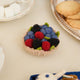Round hand-felted wool ornament designed as a mixed berry tart, topped with sparkling beaded raspberries, blackberries, and nestled blueberries. Crafted with detailed stitching and vibrant colors, it includes a hanging string and measures approximately 1.25 inches high and 2.75 inches in diameter. A whimsical and delicious-looking tribute to homemade desserts and holiday cheer