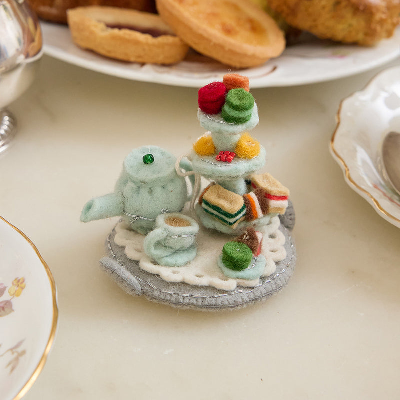 Hand-felted wool ornament featuring a pastel-blue teapot, matching teacup, and a tiered tower of colorful macarons. Embellished with embroidered details and soft textures, this miniature tea set includes a hanging string and measures approximately 2.5 inches tall. A charming nod to classic afternoon tea and vintage elegance.
