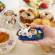 Handcrafted felt ornament of an embroidered tea tray topped with finger sandwiches, pastries, and a small bouquet of flowers. Made from merino-blend wool with soft textures and fine stitching, this ornament includes a hanging string and measures approximately 1.25 inches high and 3.75 inches wide. A charming nod to English tea parties and cozy mornings. Held in a hand for scale 