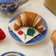 Hand-felted ornament of a golden-brown croissant resting on a pink felt napkin embroidered with a strawberry. Crafted from merino-blend wool with detailed stitching to mimic flaky layers, this ornament includes a hanging string and measures approximately 3.75 inches wide. A charming tribute to French pastry and cozy café mornings.