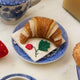 Hand-felted ornament of a golden-brown croissant resting on a pink felt napkin embroidered with a strawberry. Crafted from merino-blend wool with detailed stitching to mimic flaky layers, this ornament includes a hanging string and measures approximately 3.75 inches wide. A charming tribute to French pastry and cozy café mornings.