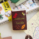 Miniature hand-sewn felt book ornament titled Nature Guide, featuring embroidered and appliquéd illustrations of plants, animals, and woodland scenes. Made from merino-blend wool with whimsical details and a hanging string. Measures approximately 2.5 inches tall by 3 inches wide. A charming tribute to the outdoors and nature lovers.