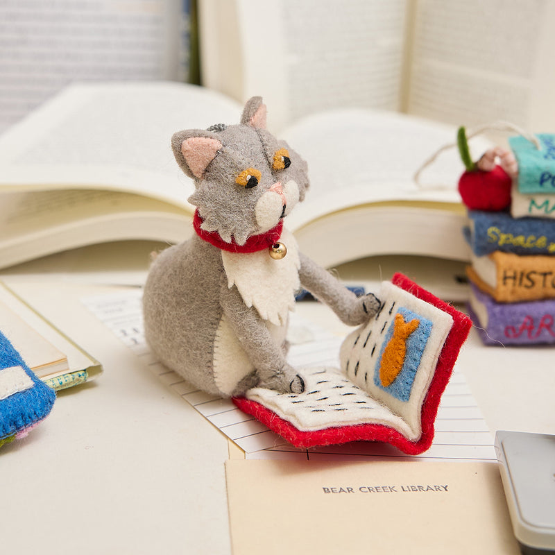 Handmade gray felted cat ornament, depicted mid-chapter in a fishy tale. The playful kitty wears a jingly collar and is fully immersed in its literary adventure. Crafted from natural wool, this whimsical ornament celebrates the love of books and cats, perfect for cat lovers, readers, and bookshop fans. Comes ready to hang with a string, adding a charming touch to any tree or reading nook.