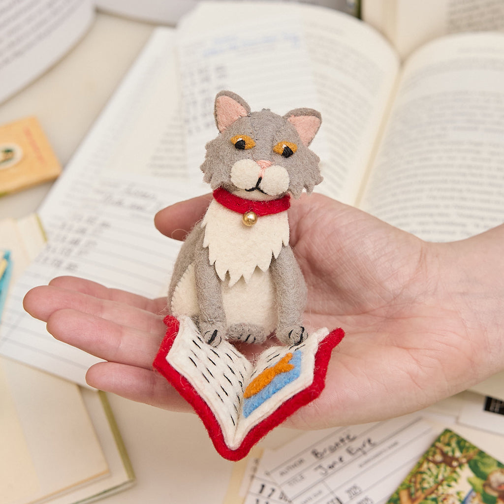 Handmade gray felted cat ornament, depicted mid-chapter in a fishy tale. The playful kitty wears a jingly collar and is fully immersed in its literary adventure. Crafted from natural wool, this whimsical ornament celebrates the love of books and cats, perfect for cat lovers, readers, and bookshop fans. Comes ready to hang with a string, adding a charming touch to any tree or reading nook. Ornament is held in  a hand to show scale.