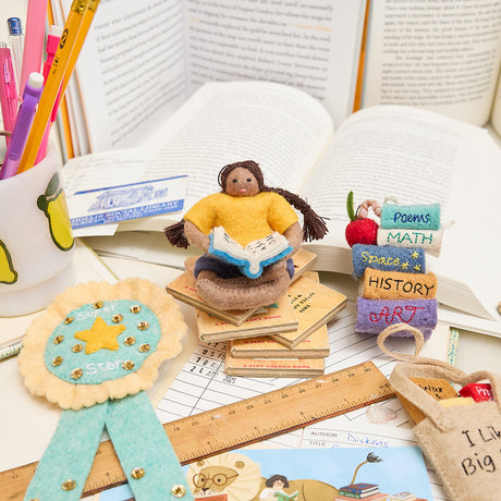 Handmade felt ornament of a young girl reading with a stack of books, super star badge and a tote of books ornaments around her. Crafted from natural merino-blended wool, her focused gaze and relaxed posture capture the magic of reading. With braided brown hair and a warm-toned complexion, this ornament adds a cozy, literary touch to reading nooks, desks, or shelves. A perfect gift for bookworms, young readers, or anyone who treasures the quiet joy of a good story. Ready to hang with a string attached
