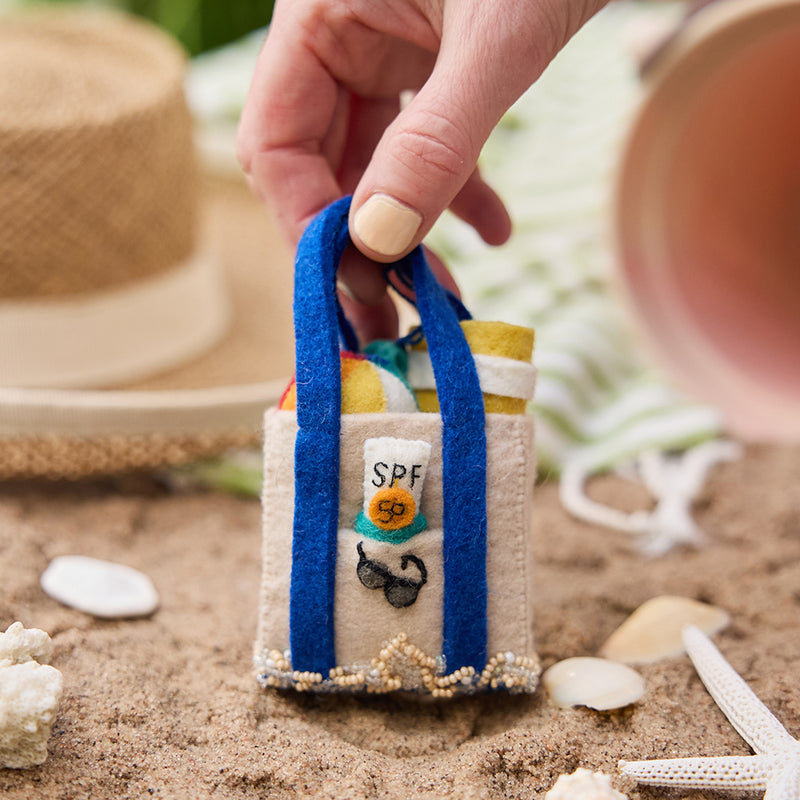 Handmade felt Beach Bag ornament from Craftspring, held by the handle over a sandy shore with seashells—heirloom-quality summer decor that brings sunny, coastal vibes to your celebrations, ethically crafted by artisans in India using sustainable wool.