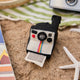 Handmade felt Polaroid Camera ornament from Craftspring, displayed on a sandy beach alongside a beach towel, book, and vintage camera—heirloom-quality summer decor that sparks memories of travel and adventure, ethically crafted by artisans in India using sustainable wool.