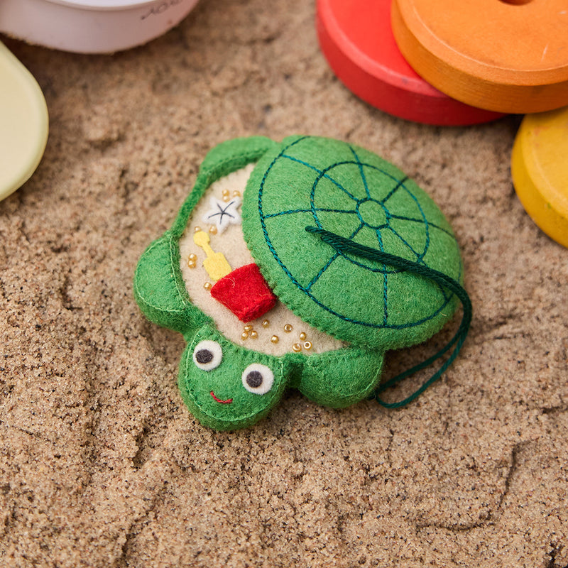 Handmade felt Turtle Sandbox ornament from Craftspring, displayed on a sandy beach with children’s beach toys—evoking nostalgic memories of childhood summers and seaside play, ethically crafted by artisans in India using sustainable wool.