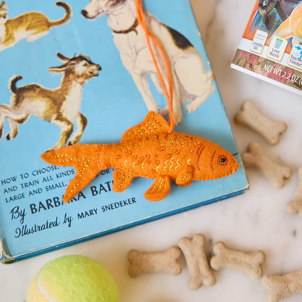Handmade felt Goldfish ornament from Craftspring, featuring intricate beaded and embroidered details—displayed on a book about pets with pet toys and treats, perfect for animal lovers and festive holiday gifting, ethically crafted by artisans using sustainable wool.
