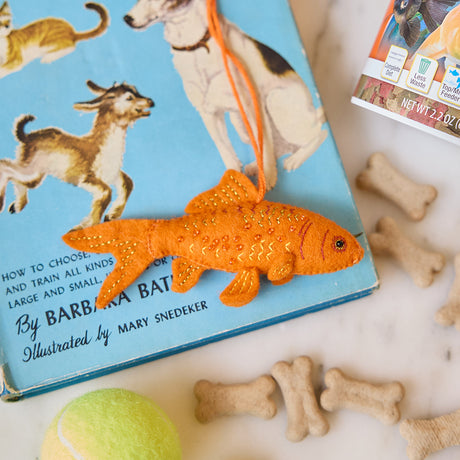 Handmade felt Goldfish ornament from Craftspring, featuring intricate beaded and embroidered details—displayed on a book about pets with pet toys and treats, perfect for animal lovers and festive holiday gifting, ethically crafted by artisans using sustainable wool.