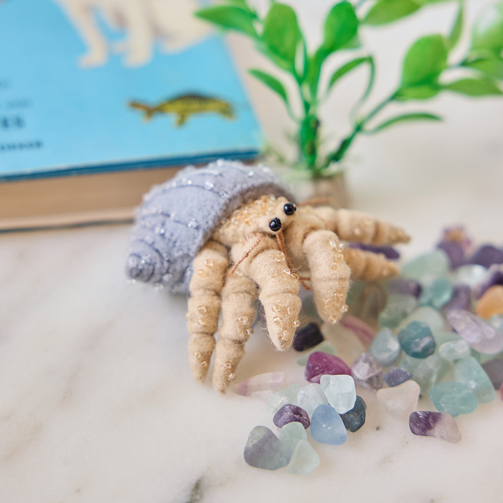 Handmade felt Hermit Crab ornament from Craftspring, featuring intricate beaded and embroidered details—displayed with aquarium rocks and a plant, perfect for ocean lovers, pet-themed decor, and whimsical holiday gifting, ethically crafted by artisans using sustainable wool.