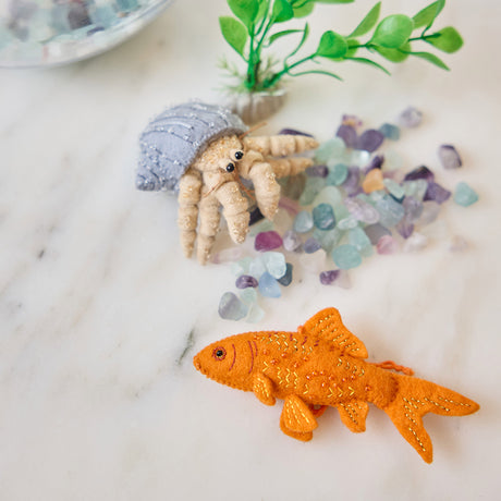 Handmade felt Goldfish and Hermit Crab ornaments from Craftspring, both featuring intricate beaded and embroidered details—displayed with aquarium rocks and a plant, perfect for pet lovers and whimsical holiday gifting, ethically crafted by artisans using sustainable wool.