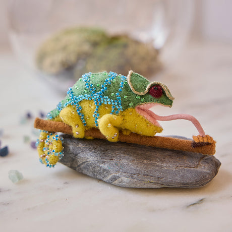 Craftspring’s handmade felt Chameleon Christmas ornament displayed on a rock—featuring beaded details, a curled tail, and a long tongue catching a bug, perfect for fun and unique holiday gifting, ethically crafted by artisans using sustainable wool.