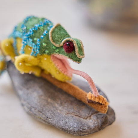 Craftspring’s handmade felt Chameleon Christmas ornament displayed on a rock—featuring beaded details, a curled tail, and a long tongue catching a bug, perfect for fun and unique holiday gifting, ethically crafted by artisans using sustainable wool.