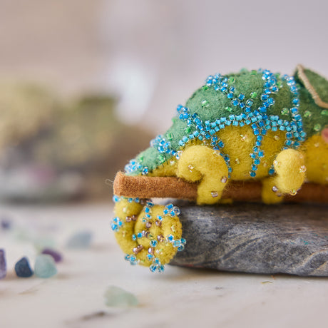 Craftspring’s handmade felt Chameleon Christmas ornament displayed on a rock—featuring beaded details, a curled tail, and a long tongue catching a bug, perfect for fun and unique holiday gifting, ethically crafted by artisans using sustainable wool.