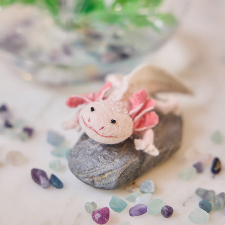 Craftspring’s handmade felt Axolotl Christmas ornament displayed among aquarium stones—featuring a cute pink design with intricate embroidered and beaded details, perfect for whimsical holiday gifting, ethically crafted by artisans using sustainable wool.