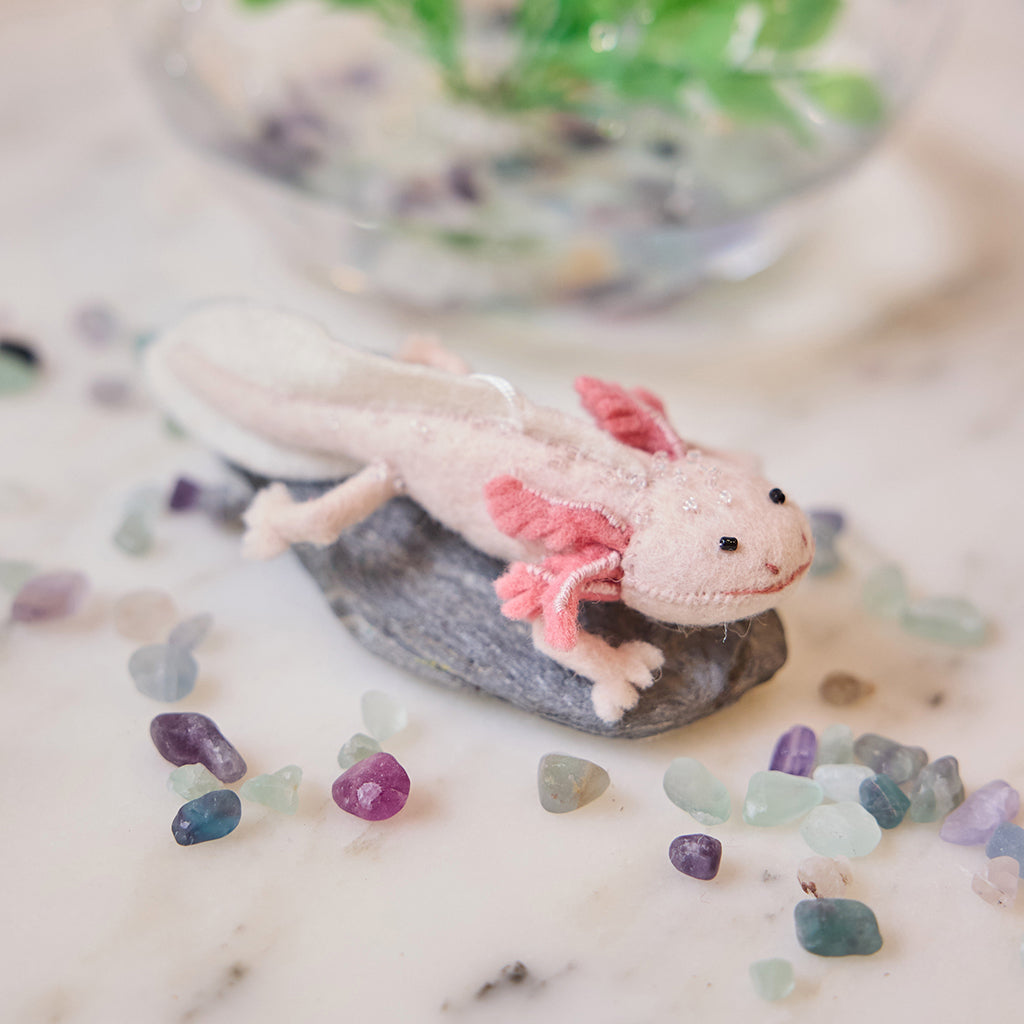 Craftspring’s handmade felt Axolotl Christmas ornament displayed among aquarium stones—featuring a cute pink design with intricate embroidered and beaded details, perfect for whimsical holiday gifting, ethically crafted by artisans using sustainable wool.
