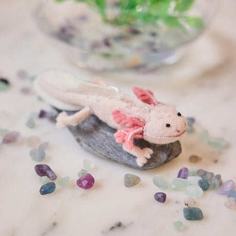 Craftspring’s handmade felt Axolotl Christmas ornament displayed among aquarium stones—featuring a cute pink design with intricate embroidered and beaded details, perfect for whimsical holiday gifting, ethically crafted by artisans using sustainable wool.