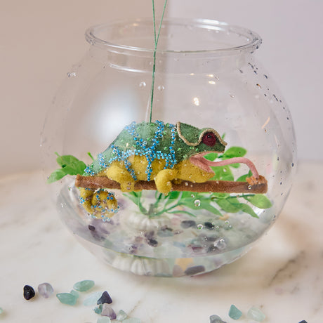 Craftspring’s handmade felt Chameleon Christmas ornament displayed inside a fish bowl with a plant—featuring beaded details, a curled tail, and a long tongue catching a bug, perfect for whimsical holiday gifting, ethically crafted by artisans using sustainable wool.
