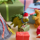 Handmade felt Santa Stegosaurus ornament from Craftspring, displayed next to a volcano school project with mini wrapped presents —featuring a green dinosaur with embroidered spots, and a red Santa hat, perfect for whimsical holiday gifting and childhood wonder, ethically crafted by artisans using sustainable wool.