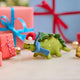 Handmade felt Santa Stegosaurus ornament from Craftspring, displayed on marble in front of festive wrapped presents—featuring a green dinosaur with embroidered spots, a red Santa hat, and a blue scarf, ethically crafted by artisans using sustainable wool for cheerful holiday decor.