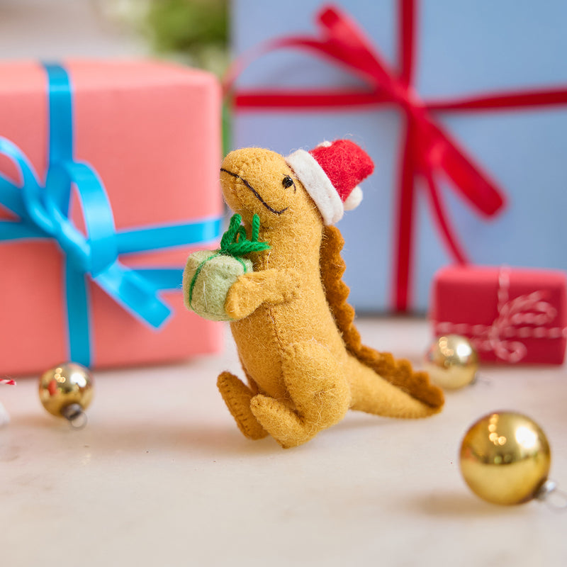 Handmade felt Orange T-Rex Dinoclaus ornament from Craftspring, displayed on marble in front of festive wrapped presents—featuring a cheerful orange dinosaur with a Santa hat and green scarf, perfect for dino-loving kids and joyful holiday gifting, ethically crafted by artisans using sustainable wool.