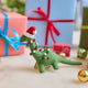 Handmade felt Merry & Bright Santa Brontosaurus ornament from Craftspring, displayed in front of festive wrapped presents—featuring a green dinosaur with a red Santa hat and a garland wrapped around its long neck, perfect for dino-loving kids and joyful holiday gifting, ethically crafted by artisans using sustainable wool.