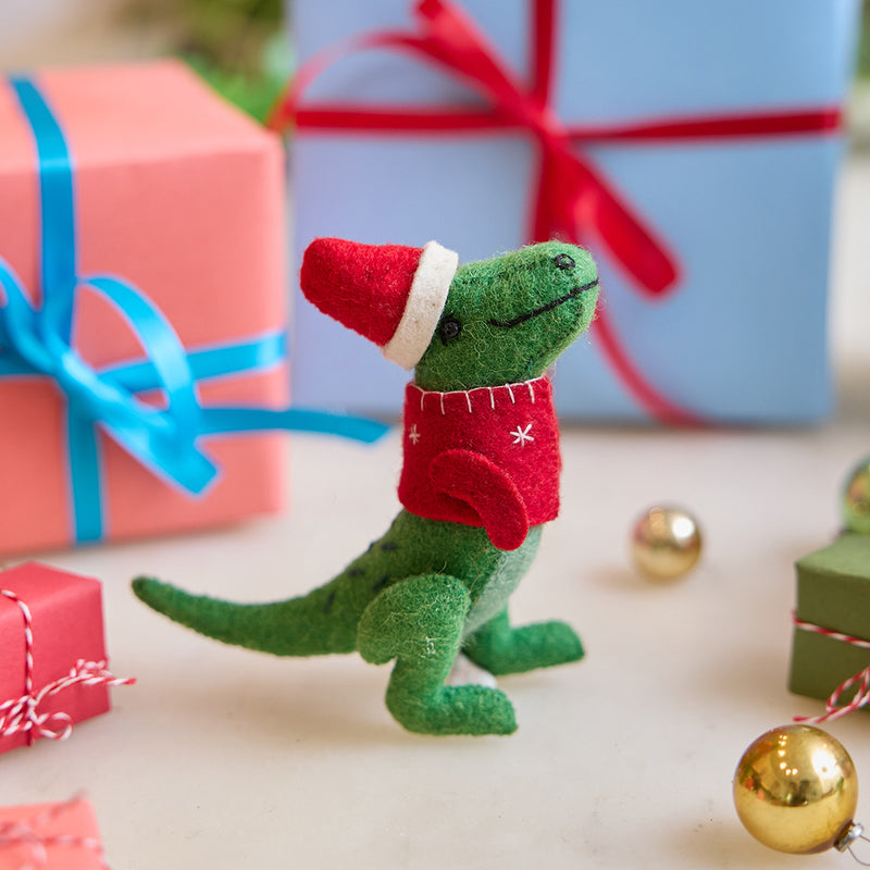 Handmade felt Santasaurus T-Rex ornament from Craftspring, displayed in front of festive wrapped presents—perfect for dino-loving kids and joyful holiday gifting, featuring a red Santa hat, cozy red sweater, and embroidered details, ethically crafted using sustainable wool.