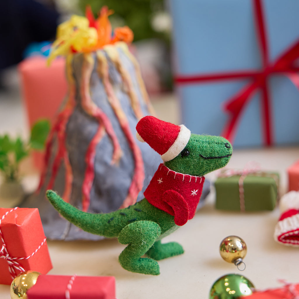 Handmade felt Santasaurus T-Rex ornament from Craftspring, displayed in front of a volcano school project with wrapped presents nearby—featuring a red Santa hat, red sweater, and embroidered details, perfect for dino-loving kids and festive holiday gifting, ethically crafted using sustainable wool.