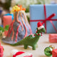 Handmade felt Merry & Bright Santa Brontosaurus ornament from Craftspring, displayed in front of a volcano school project with wrapped presents nearby—featuring a green dinosaur with a red Santa hat and a festive garland, perfect for dino-loving kids and playful holiday gifting, ethically crafted by artisans using sustainable wool.