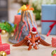Handmade felt Merry Pterodactyl ornament from Craftspring, displayed in front of a volcano school project with mini wrapped presents nearby—perfect for dino-loving kids and festive holiday gifting, featuring embroidered details, a Santa hat, and a “MERRY” garland, ethically crafted using sustainable wool.