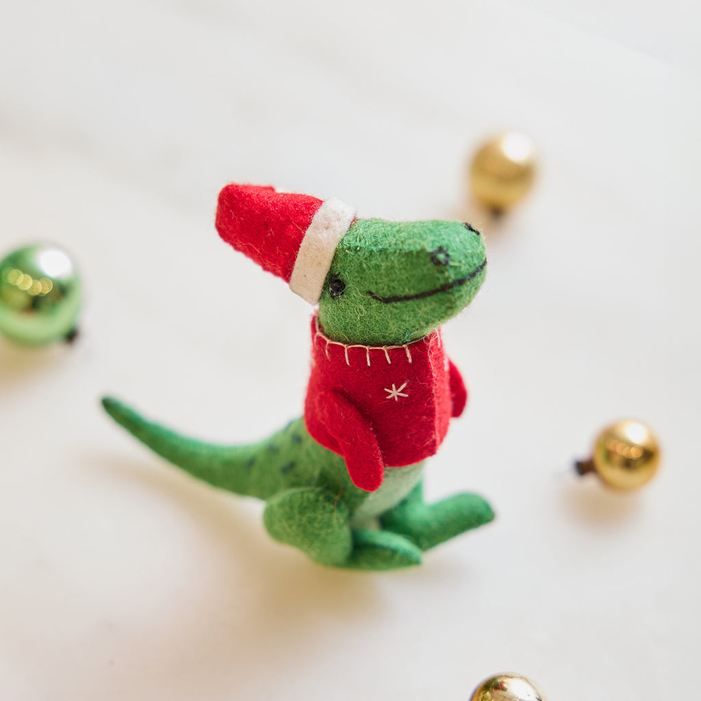 Handmade felt Santasaurus T-Rex ornament from Craftspring, displayed on marble with festive bobble ornaments—featuring a red Santa hat, red sweater, and embroidered details, perfect for dino-loving kids and cheerful holiday gifting, ethically crafted using sustainable wool.