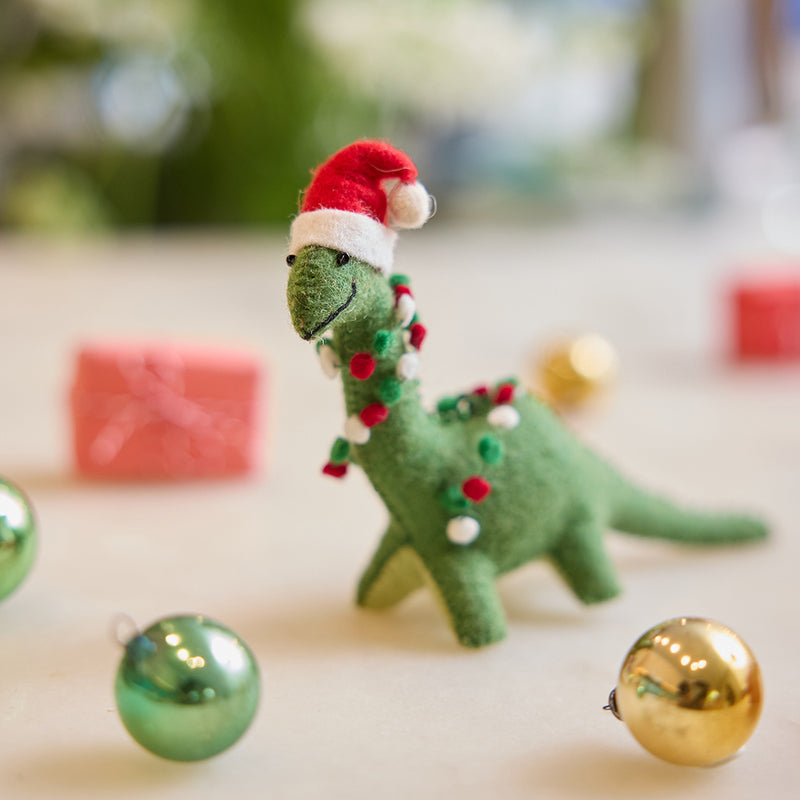 Handmade felt Merry & Bright Santa Brontosaurus ornament from Craftspring, displayed on marble with mini wrapped presents and bobble ornaments—featuring a green dinosaur with a festive red Santa hat, perfect for joyful holiday gifting and playful seasonal decor, ethically crafted by artisans using sustainable wool.