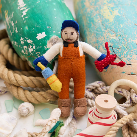 Lifestyle photo of Craftspring’s handmade felt Maine Lobsterman Christmas ornament, featuring beaded and embroidered details—holding a bright red lobster and buoy, displayed with nautical props for a festive coastal holiday feel. Ethically crafted by artisans using sustainable wool.