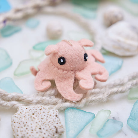 Handmade felt Dumbo Octopus ornament from Craftspring, featuring adorable curly pink tentacles and charming embroidered details—cuddled among sea glass, rope, and shells for a sweet coastal Christmas vibe, lovingly crafted by artisans using sustainable wool.