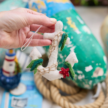 Handmade felt Anchor ornament from Craftspring, featuring beaded barnacles, appliqué seaweed and starfish, and draped in netting—held gently in hand against a nautical setting with rope and lobster buoys, perfect for holiday gifting and sustainably handmade by artisans using wool.
