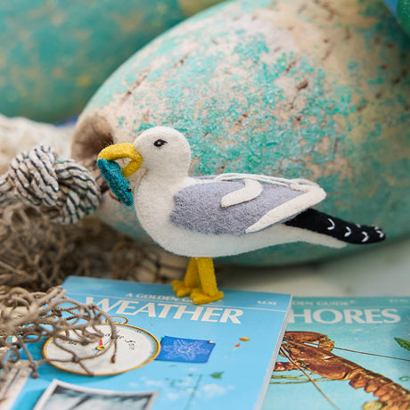 Handmade felt Seagull ornament from Craftspring, featuring embroidered details and holding a fish in its beak—displayed with lobster buoys and netting, perfect for nautical Christmas decor and festive gifting, sustainably handmade by artisans using wool.