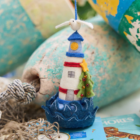 Handmade felt Holiday Lighthouse Christmas ornament from Craftspring, featuring a seagull on top, crashing waves at the base, and a Christmas tree on the side—adorned with intricate beaded and embroidered details, displayed with lobster buoys and netting for a festive nautical holiday vibe, ethically crafted by artisans using sustainable wool.
