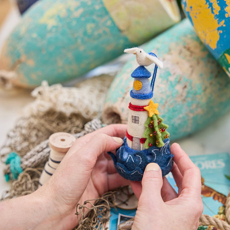 Handmade felt Holiday Lighthouse ornament from Craftspring, featuring a seagull on top, crashing waves, and a Christmas tree—held gently in hands against a nautical backdrop with lobster buoys and netting, perfect for festive holiday gifting, ethically crafted by artisans using sustainable wool.