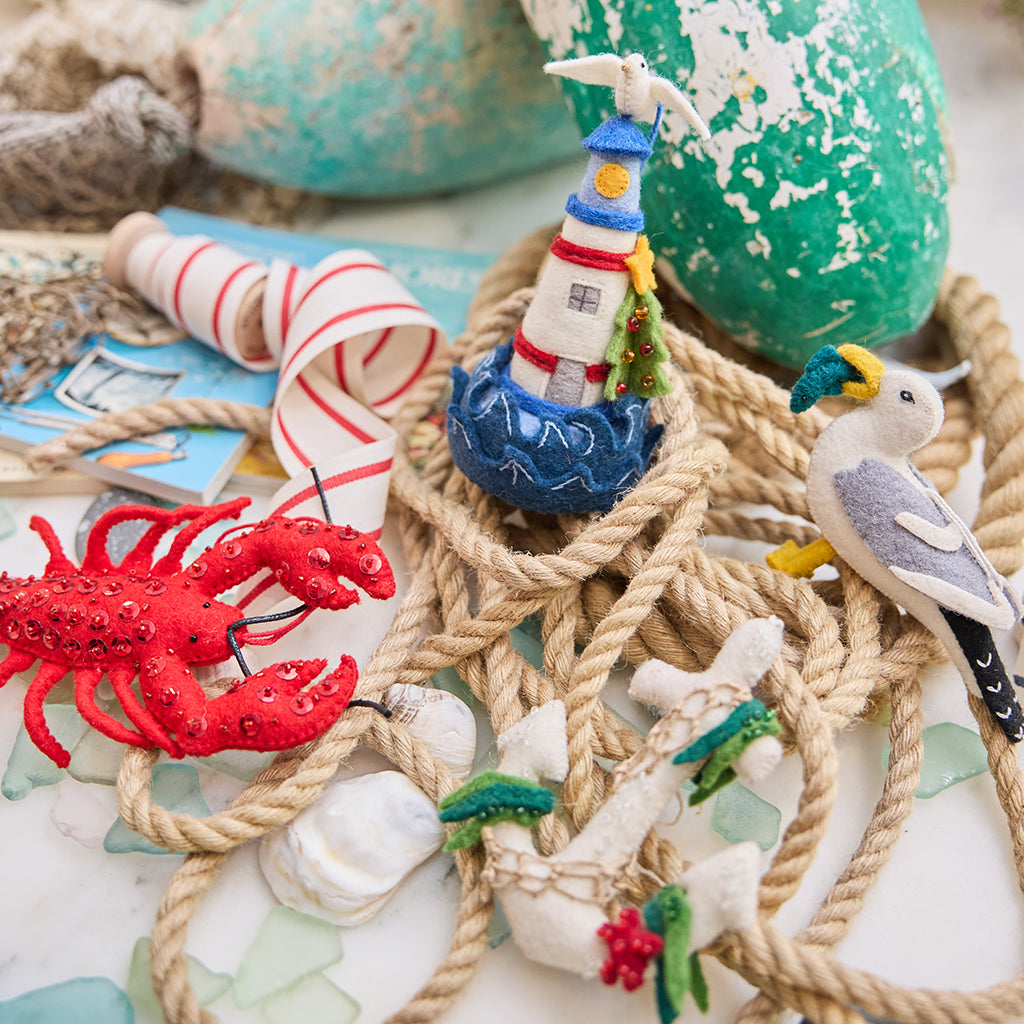 Handmade felt Anchor, Seagull, Holiday Lighthouse, and Lobster ornaments from Craftspring, featuring detailed embroidery, appliqué, and beaded accents—displayed with rope and lobster buoys, perfect for nautical holiday decor and gifting, sustainably handmade by artisans using wool.