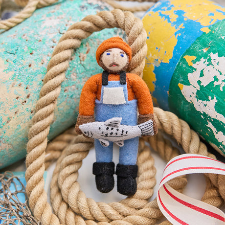 Handmade felt Old Man Salmon ornament from Craftspring, featuring embroidered details, a fishing bib, and a proudly held salmon—displayed with nautical rope, lobster buoys, and netting for a whimsical touch of maritime Christmas cheer, sustainably handmade by artisans using wool.