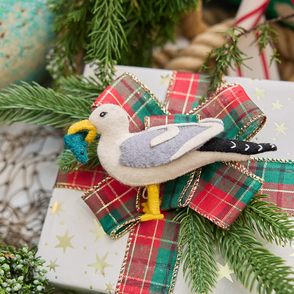 Handmade felt Seagull ornament from Craftspring, featuring embroidered details and holding a fish in its beak—displayed on a holiday gift, perfect for nautical Christmas decor, sustainably handmade by artisans using wool.