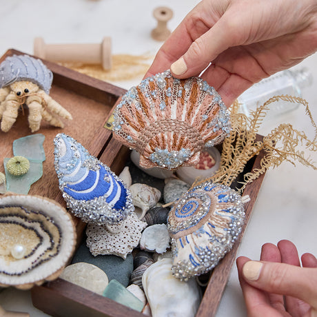 Craftspring’s Mussel Shell, Oyster, Moon Snail Shell, Scallop Shell, and Hermit Crab ornaments — all intricately beaded and embroidered, the shells made of satin and the Oyster and Hermit Crab made of felt — displayed in a box of ocean treasures with seashells and sea glass, creating a playful and festive holiday scene, ethically crafted by artisans.