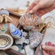Craftspring’s Mussel Shell, Oyster, Moon Snail Shell, Scallop Shell, and Hermit Crab ornaments — all intricately beaded and embroidered, the shells made of satin and the Oyster and Hermit Crab made of felt — displayed in a box of ocean treasures with seashells and sea glass, creating a playful and festive holiday scene, ethically crafted by artisans.