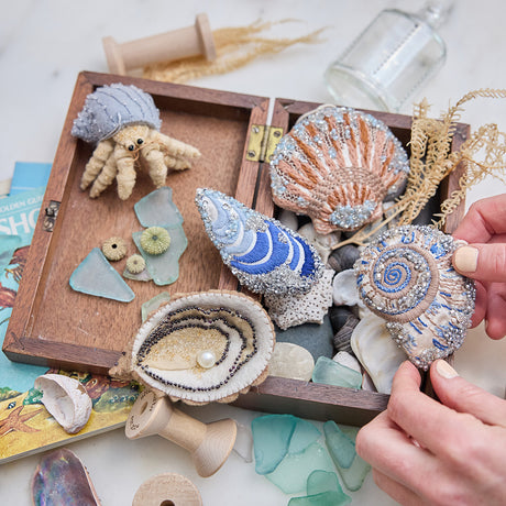 Craftspring’s Mussel Shell, Oyster, Moon Snail Shell, Scallop Shell, and Hermit Crab ornaments — all intricately beaded and embroidered, the shells made of satin and the hermit crab made of felt — displayed in a box of ocean treasures with seashells and sea glass, creating a playful and festive holiday scene, ethically crafted by artisans.
