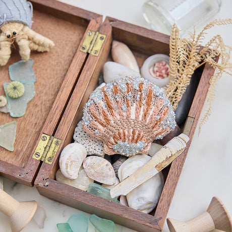 Craftspring’s Scallop Shell Ornament — made of satin with intricate beaded and embroidered details — displayed in a box of ocean treasures with seashells and sea glass, creating a playful and festive holiday scene, ethically crafted by artisans.