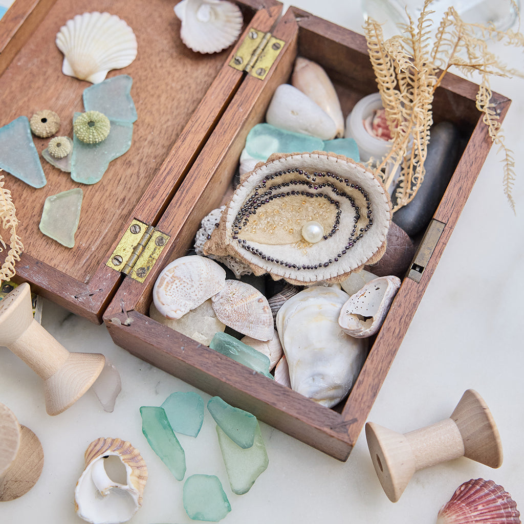 Lifestyle photo of Craftspring’s handmade felt Oyster Christmas ornament, nestled in a wooden box filled with shells and sea glass—featuring a beaded pearl and shimmering embroidered details, ethically crafted by artisans using sustainable wool for ocean-inspired holiday charm.









