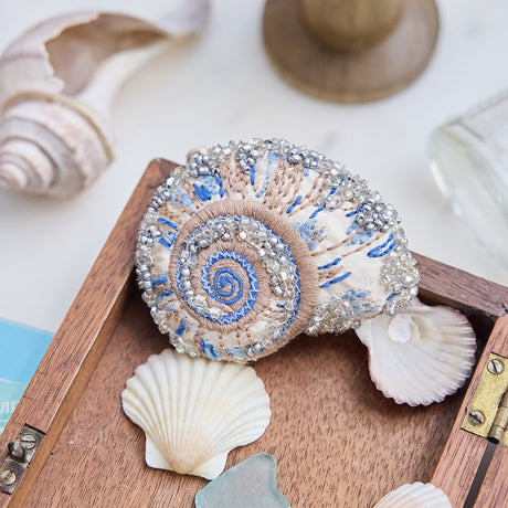 Craftspring’s Moon Snail Shell Ornament — made of satin with intricate beaded and embroidered details — displayed in a box of ocean treasures with seashells and sea glass, creating a playful and festive holiday scene, ethically crafted by artisans.
