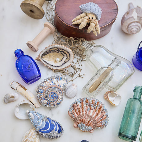Craftspring’s Mussel Shell, Oyster, Moon Snail Shell, Scallop Shell, and Hermit Crab ornaments — all intricately beaded and embroidered, the shells made of satin and the Oyster and Hermit Crab made of felt — displayed among seashells and glass bottles, creating a festive coastal holiday scene, ethically crafted by artisans.